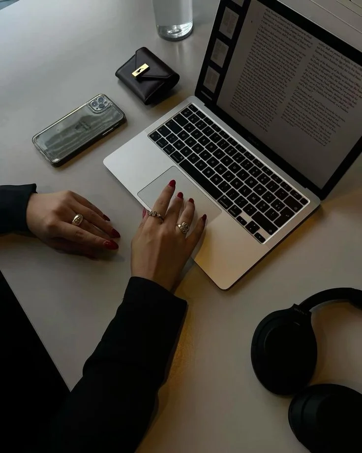 A woman’s hands working on a laptop at a desk, beside a phone and headphones